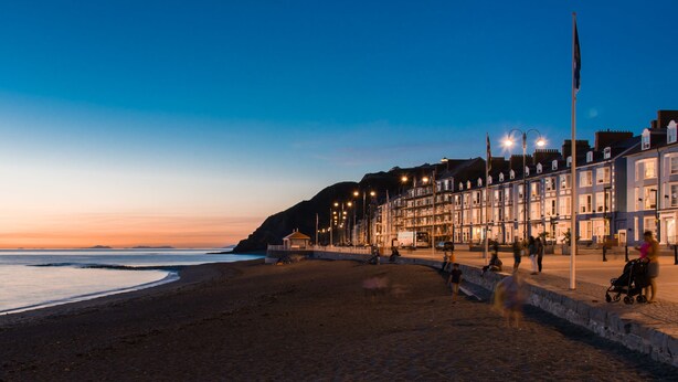 A coastal promenade with buildings along the shore, a sandy beach, and a calm sea at dusk. - Gwesty'r Marine Hotel & Spa (Aberystwyth)