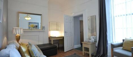 Una sala de estar acogedora con un sofá, una mesa de centro y una lámpara. Hay una puerta que da a otra habitación y una ventana con cortinas.