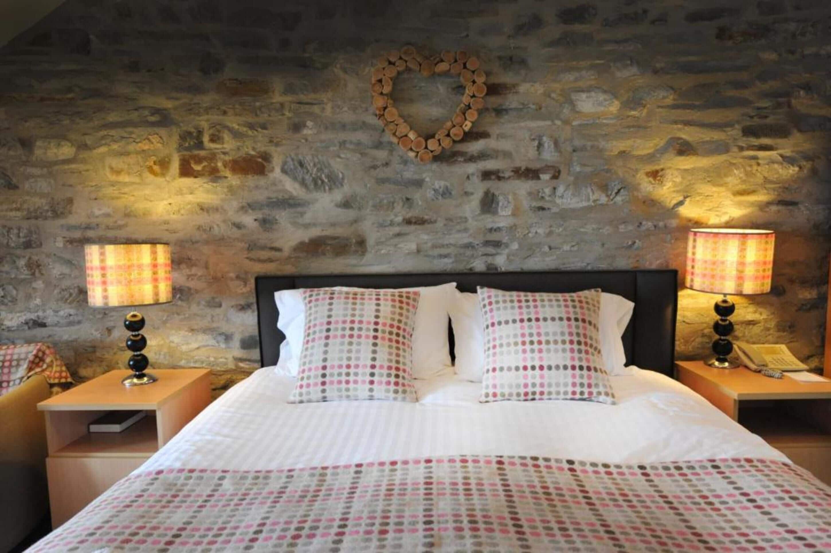 Un dormitorio con pared de piedra, una decoración con forma de corazón, dos lámparas de noche y una cama con ropa de cama estampada.