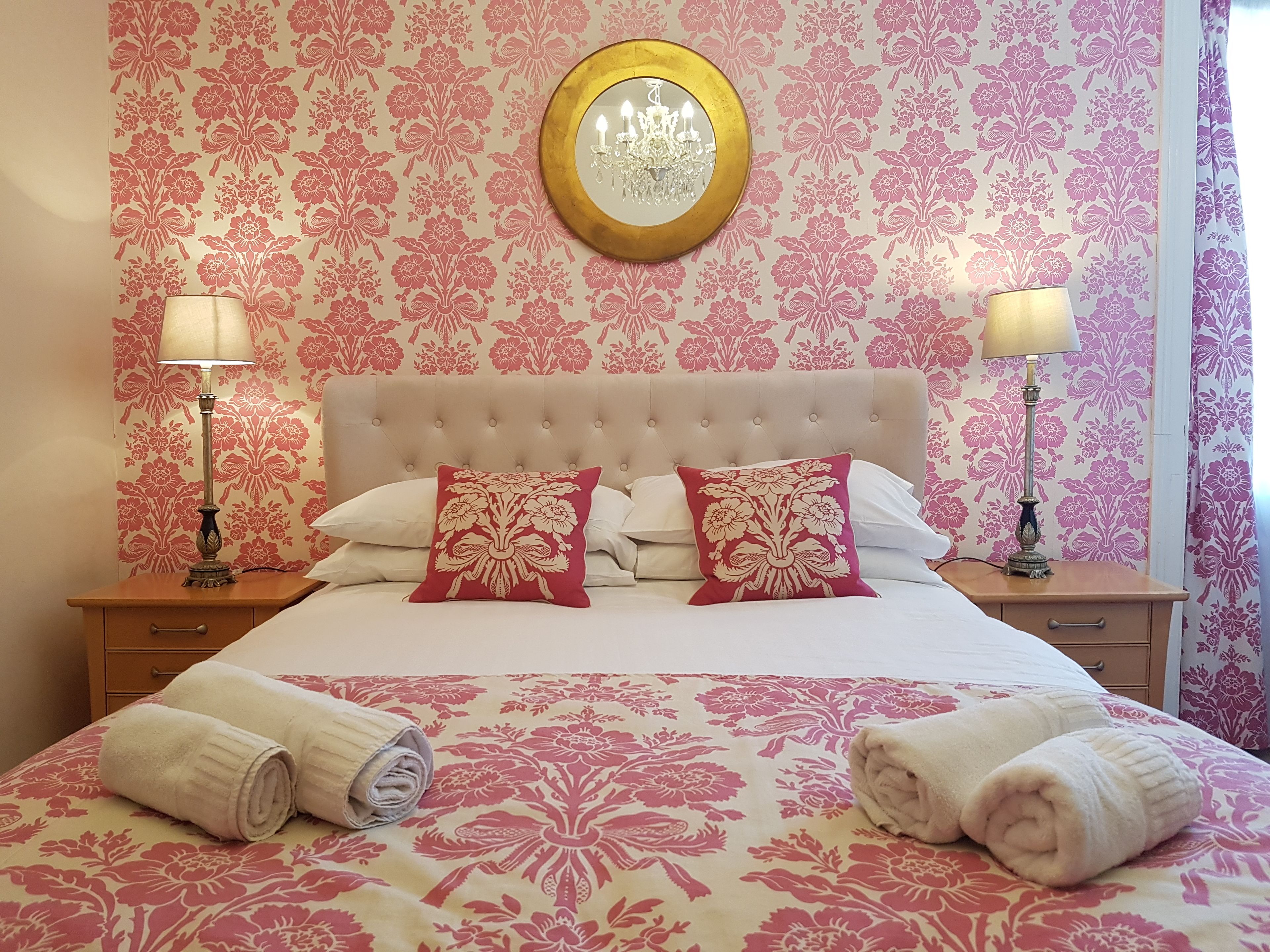A neatly made bed with floral-patterned bedding, two bedside lamps, and a round mirror above the headboard.