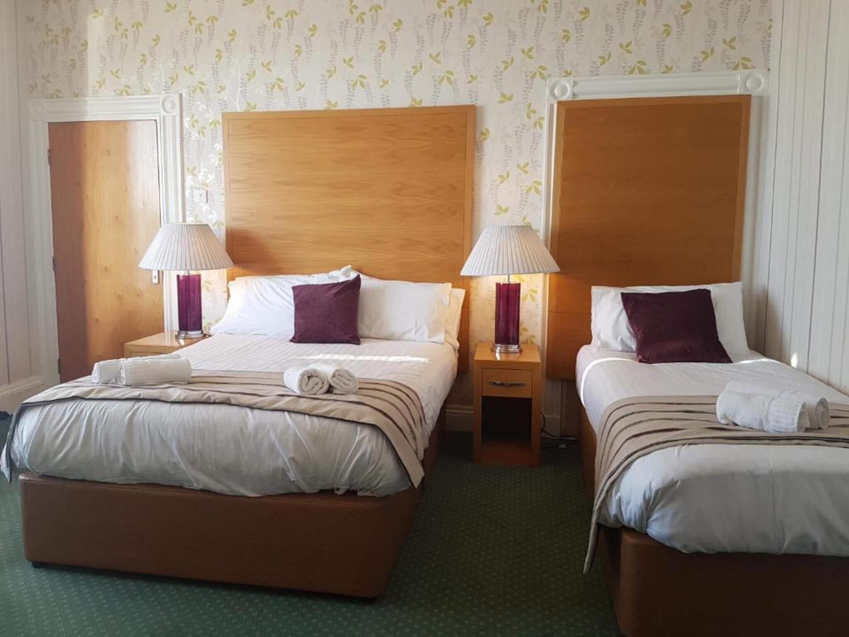 A hotel room with two beds, each with a wooden headboard, white bedding, and purple pillows. There are two bedside lamps and a small wooden nightstand between the beds.