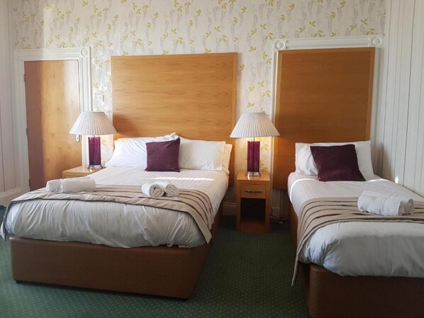 Habitación de hotel con dos camas, cada una con cabecera de madera, ropa de cama blanca y almohadas moradas. Hay dos lámparas de noche y una mesita de noche de madera entre las camas.