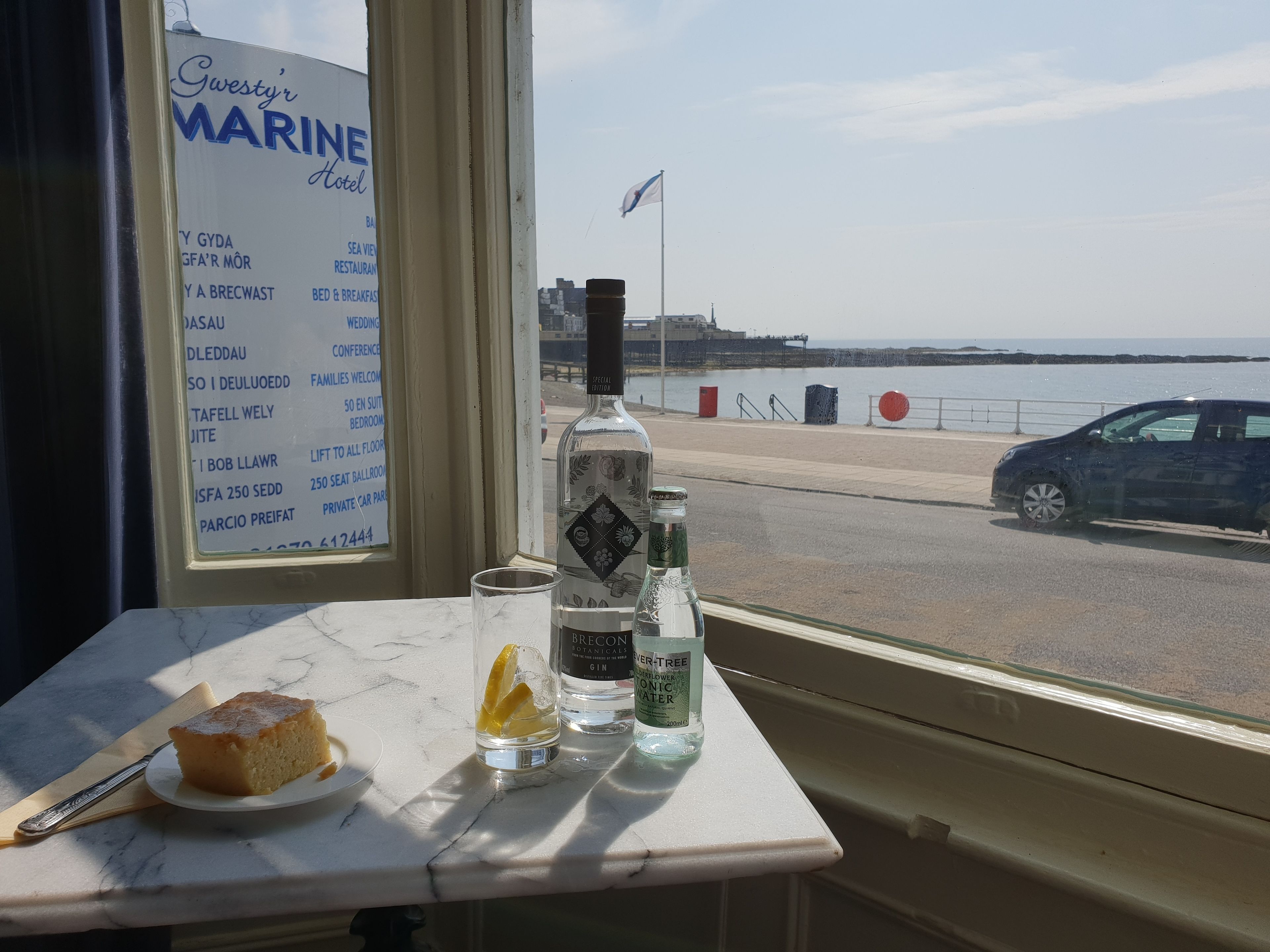 A table with a slice of cake, a glass with a lemon slice, and a bottle of gin, with a view of a coastal road and a flag in the background.
