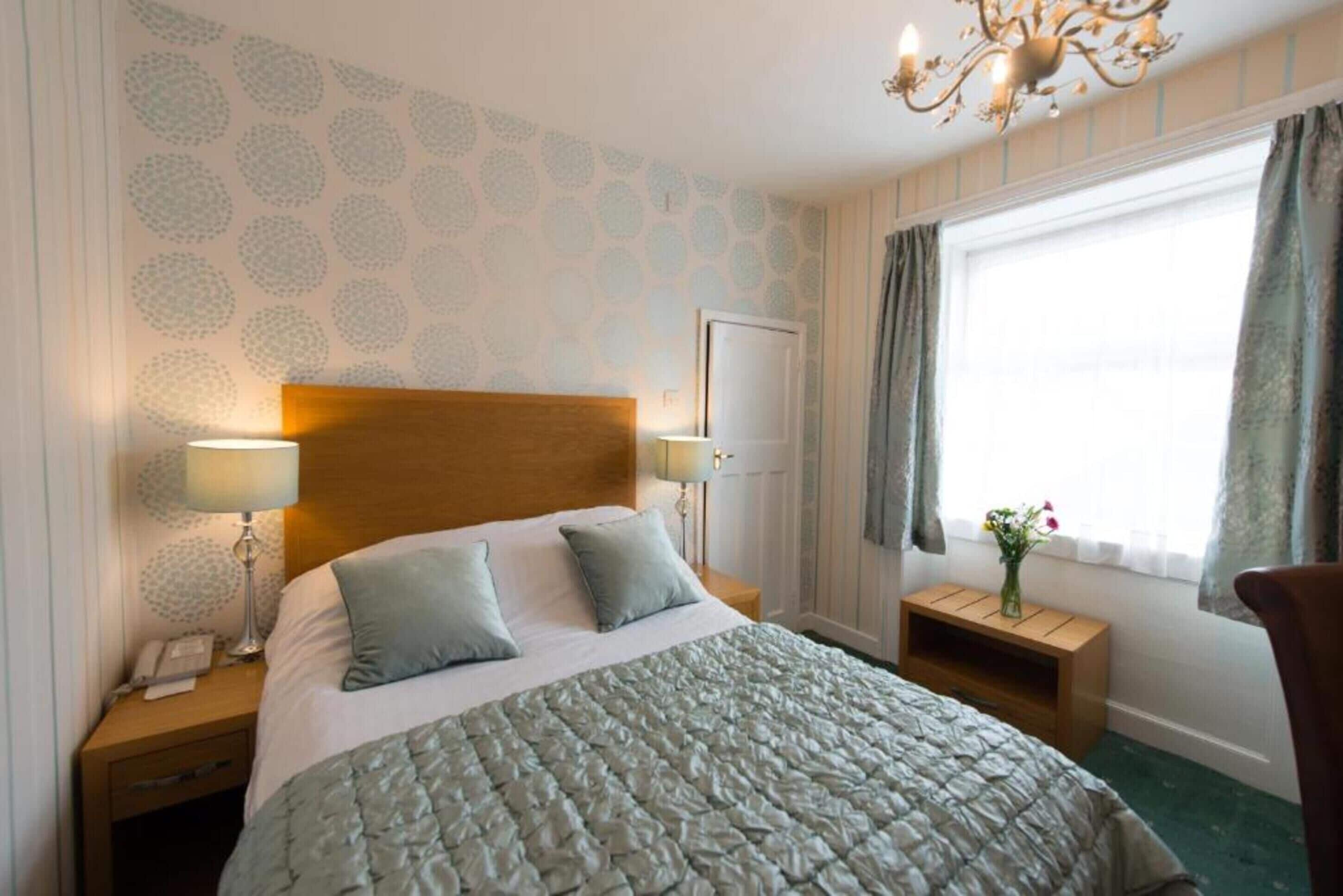 A hotel room with a large bed, two bedside tables with lamps, a wooden headboard, a window with curtains, and a chandelier.