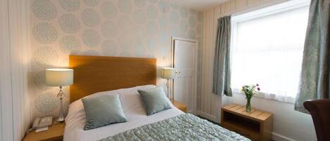 A hotel room with a large bed, two bedside tables with lamps, a wooden headboard, a window with curtains, and a chandelier.