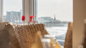 A view from a window overlooking a pier and buildings, with a blurred foreground featuring a wicker chair and a vase with flowers. - Gwesty'r Marine Hotel & Spa (Aberystwyth)