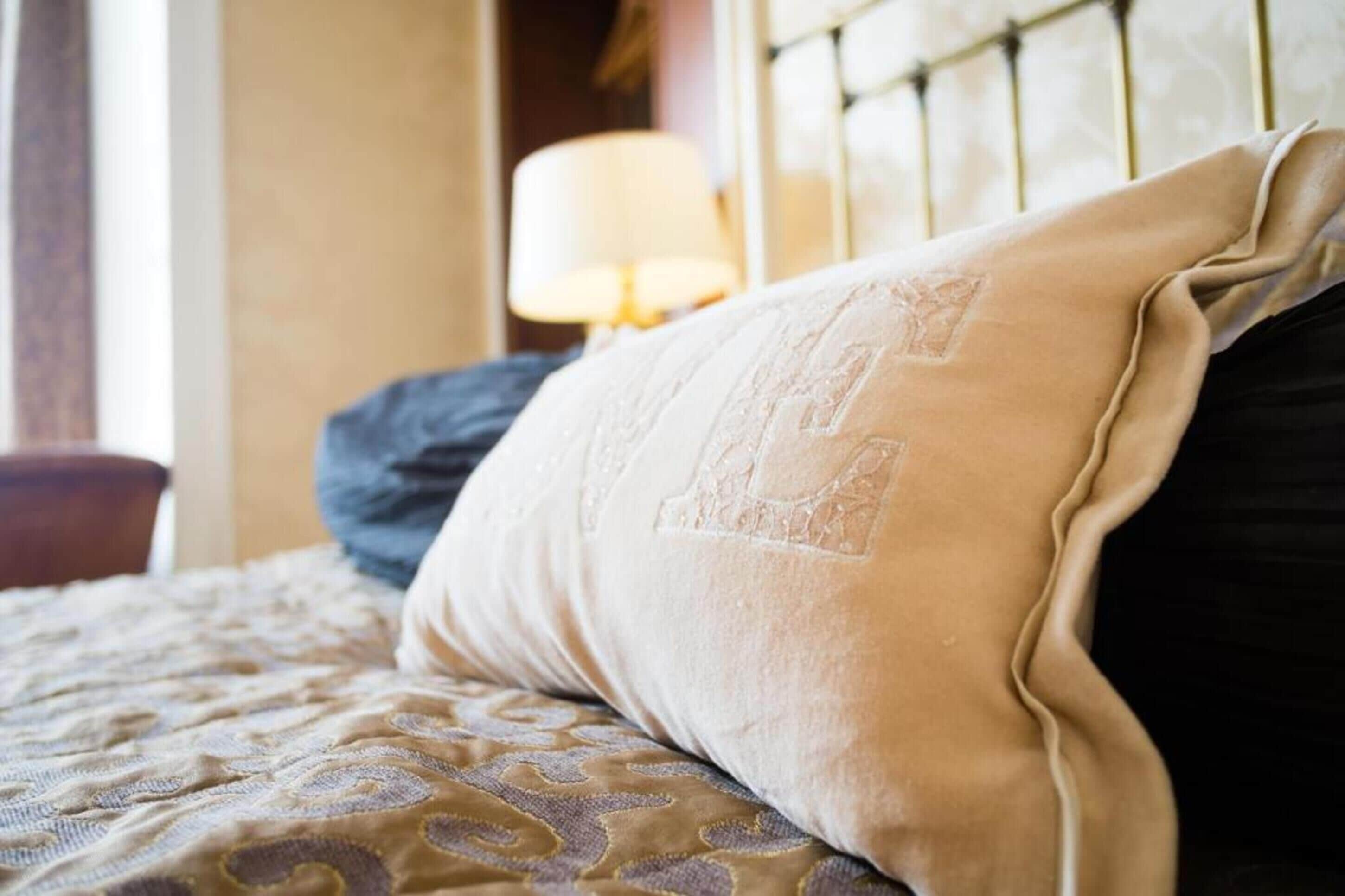 A bed with a patterned bedspread, a beige pillow with a large embroidered letter, and a dark-colored pillow.