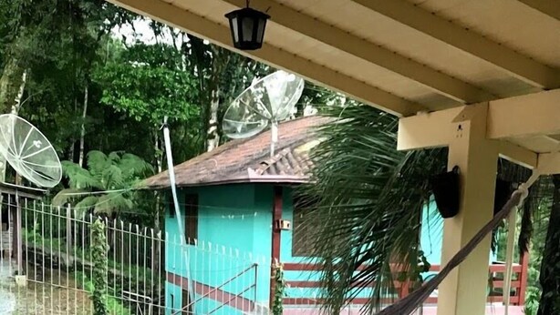 Una hamaca en un patio con baldosas, una mecedora de madera, una casa pequeña al fondo y una cerca de metal.