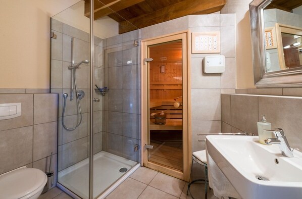 A bathroom with a glass shower enclosure, a wooden sauna door, and a white sink with a mirror above it.
