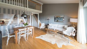 A modern living space with a wooden dining table, white chairs, a white sofa, and a white armchair. A staircase leads to an upper level. - Lerchenhof Reestow (Rankwitz)