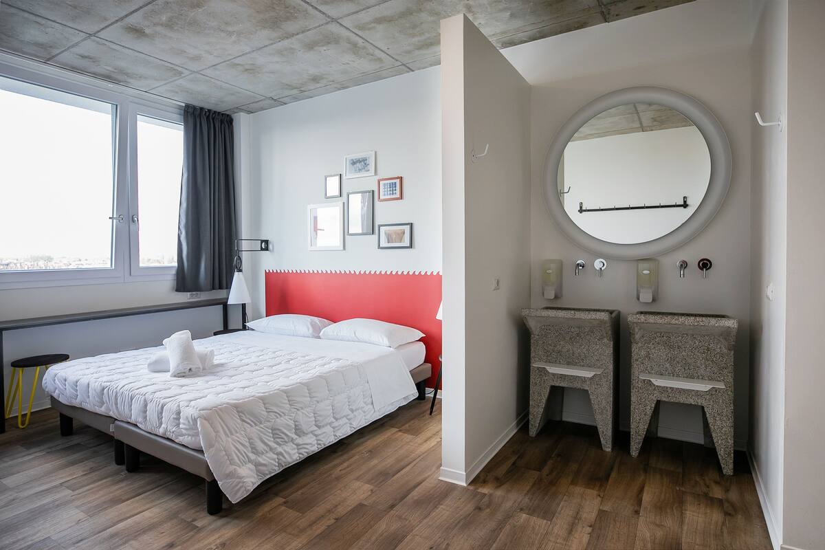 A modern bedroom with a large bed, a round mirror, two bedside tables, and a window with curtains.