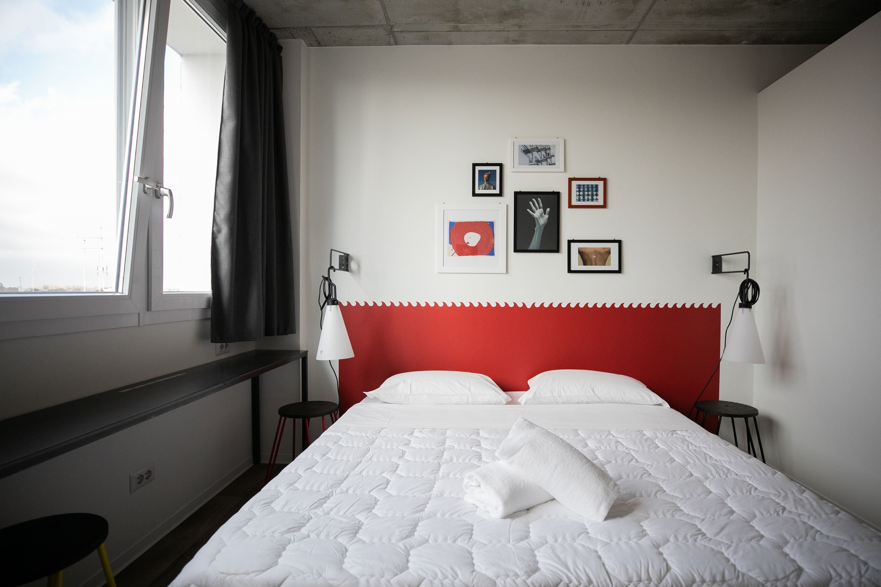 Una habitación de hotel con cama, cabecera roja, ventana con cortinas, escritorio y silla.