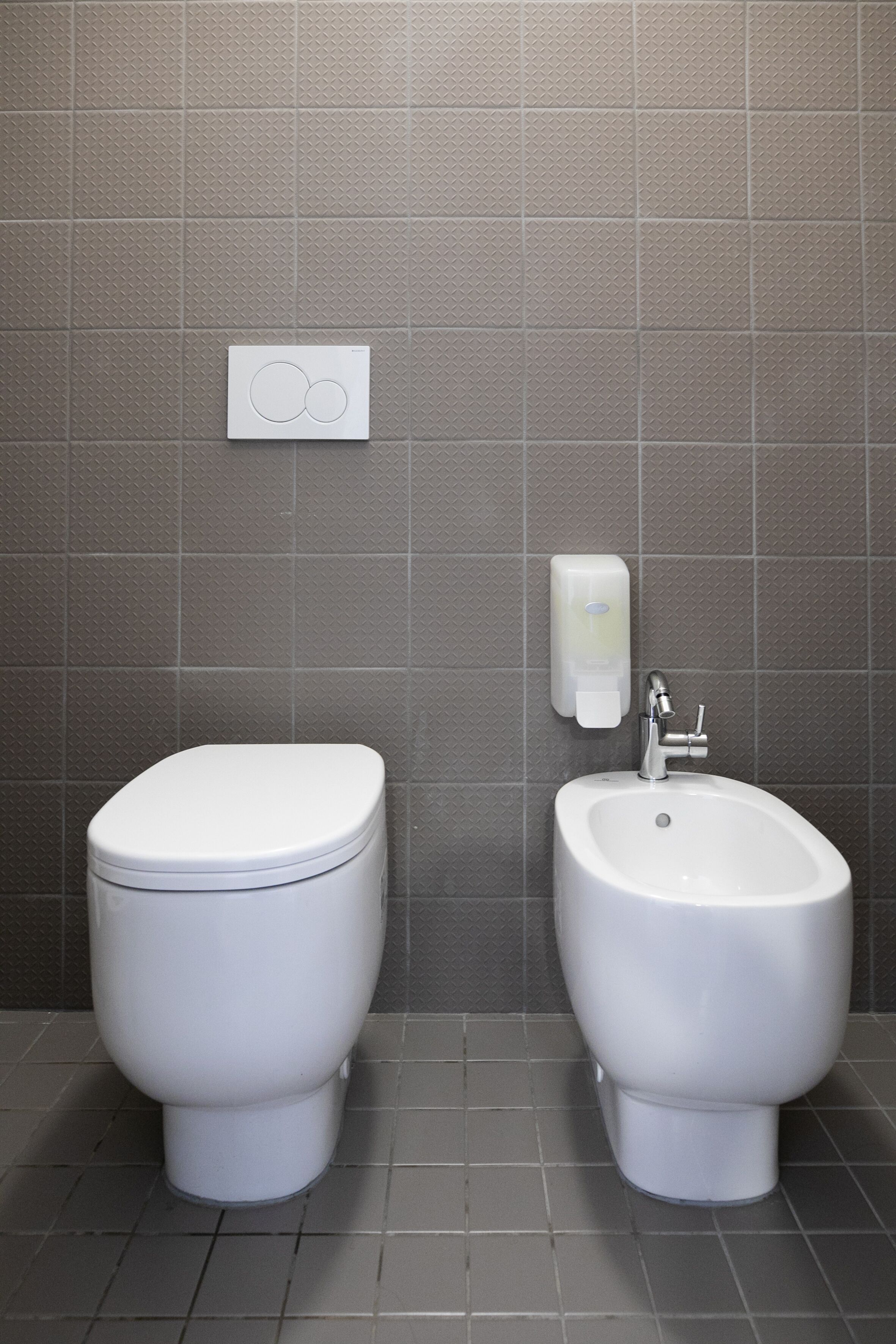 Een badkamer met toilet en bidet, wandtegels in grijstinten, en een zeepdispenser.