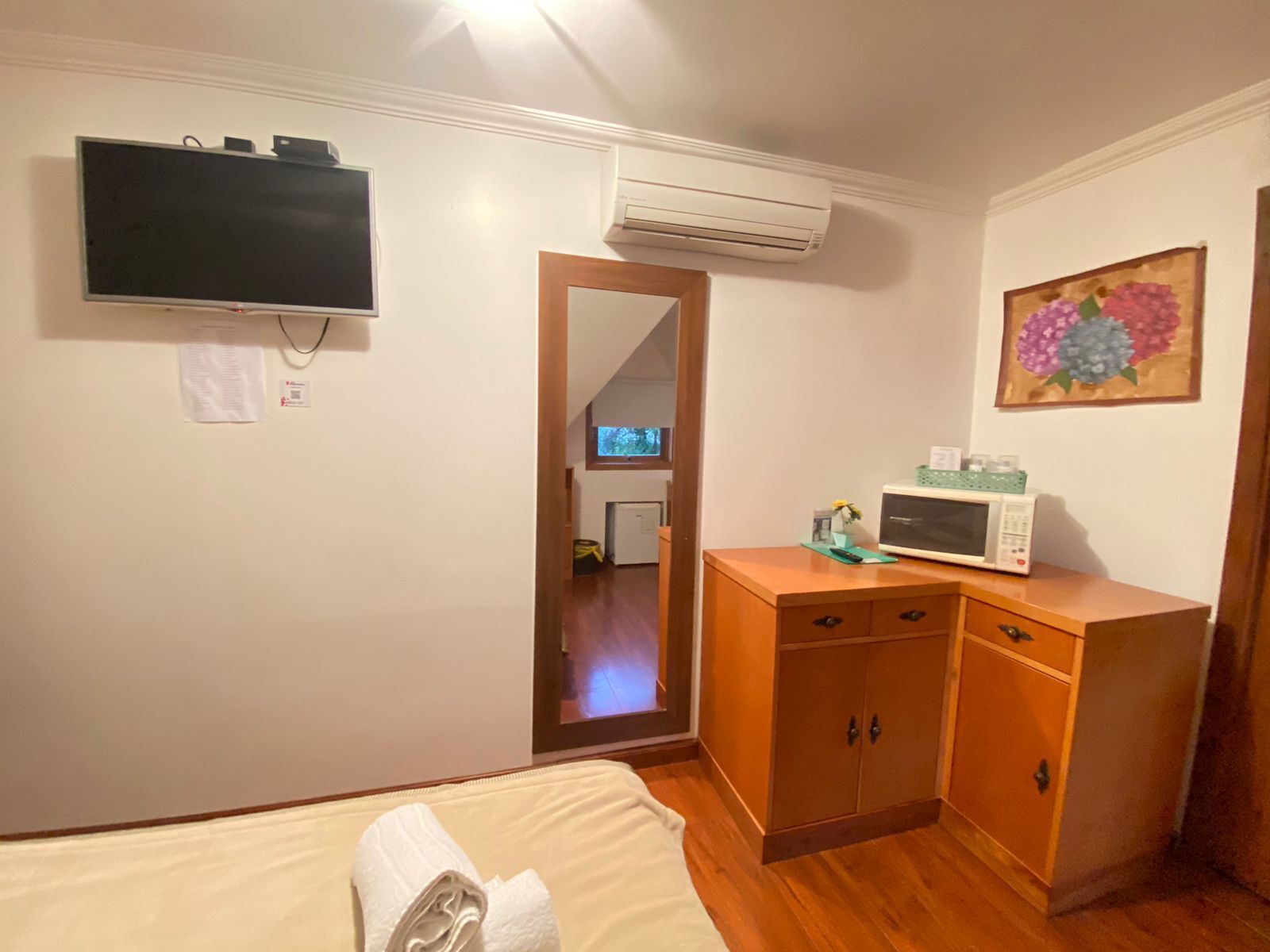 Habitación de hotel con cama, un armario de madera, microondas, unidad de aire acondicionado, televisor y un cuadro en la pared.