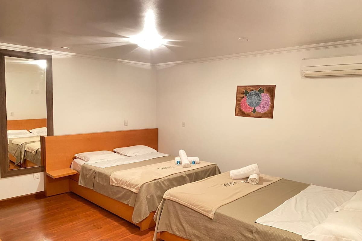 A hotel room with two single beds, a wooden headboard, a small table with toiletries, a mirror, and a wall-mounted air conditioner.