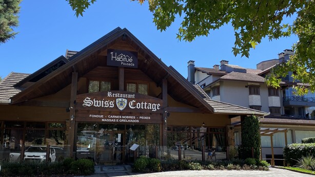 A wooden building with a sign reading "Habitué Pousada Restaurant Swiss Cottage" and "FONDUE - CARNES NOBRES - PEIXES MASSAS E GRELHADOS." - Pousada Habitue (Gramado)