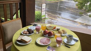 A table set with breakfast items including bread, ham, eggs, and fruit, with a view of a wet street and greenery outside. - Pousada Habitue (Gramado)