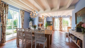 A spacious dining area with a wooden table and white chairs, a chandelier, and large windows with floral curtains. - Reetdachhaus in Krienke (Rankwitz)