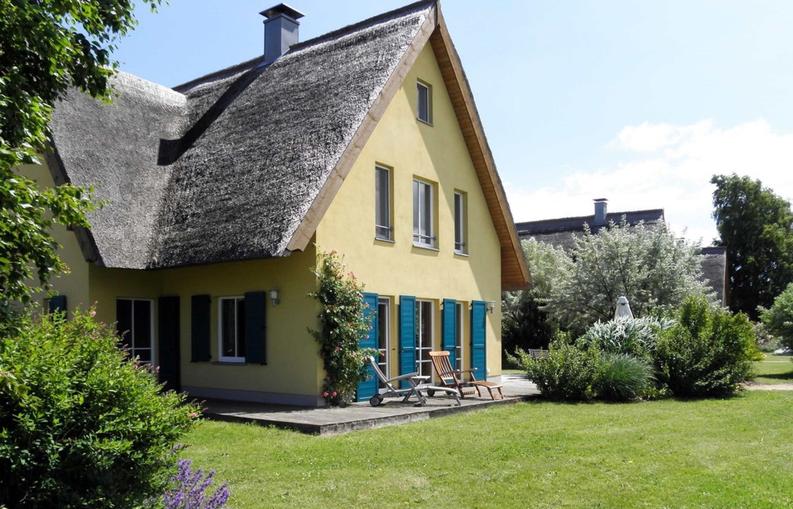 A yellow house with a thatched roof, blue shutters, and a garden with a lawn and shrubs.