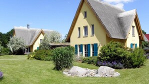 A yellow building with a steeply pitched roof and blue shutters, surrounded by a well-maintained garden with rocks and shrubs. - Reetdachhaus in Krienke (Rankwitz)