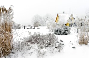 Paisaje nevado con una casa amarilla, un banco y vegetación escasa.