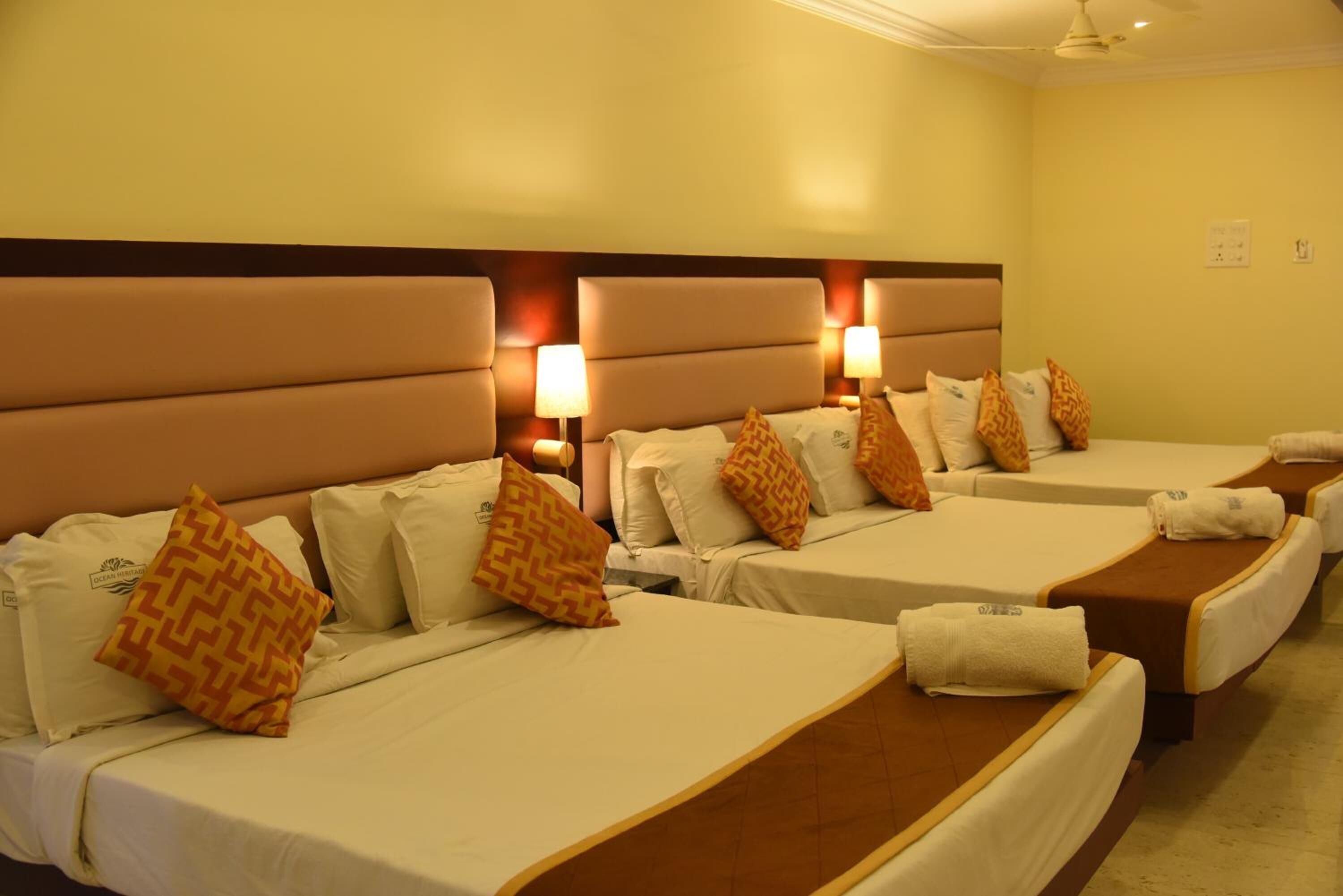 Habitación de hotel con cuatro camas dispuestas en fila, cada una con cabecera, almohadas y mesita de noche.