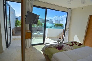 A hotel room with a bed, a flat-screen TV, a sliding glass door to a balcony, and a view of the surrounding buildings.