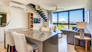 Una sala de estar moderna con una gran mesa de comedor, sillas blancas, un sofá y una mesa de centro. Se observa una escalera con barandilla negra y un televisor montado en la pared.