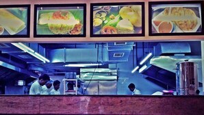 A kitchen area with stainless steel appliances, a marble countertop, and framed food menus on the wall. - Hotel mookambika palace (Kundapur)