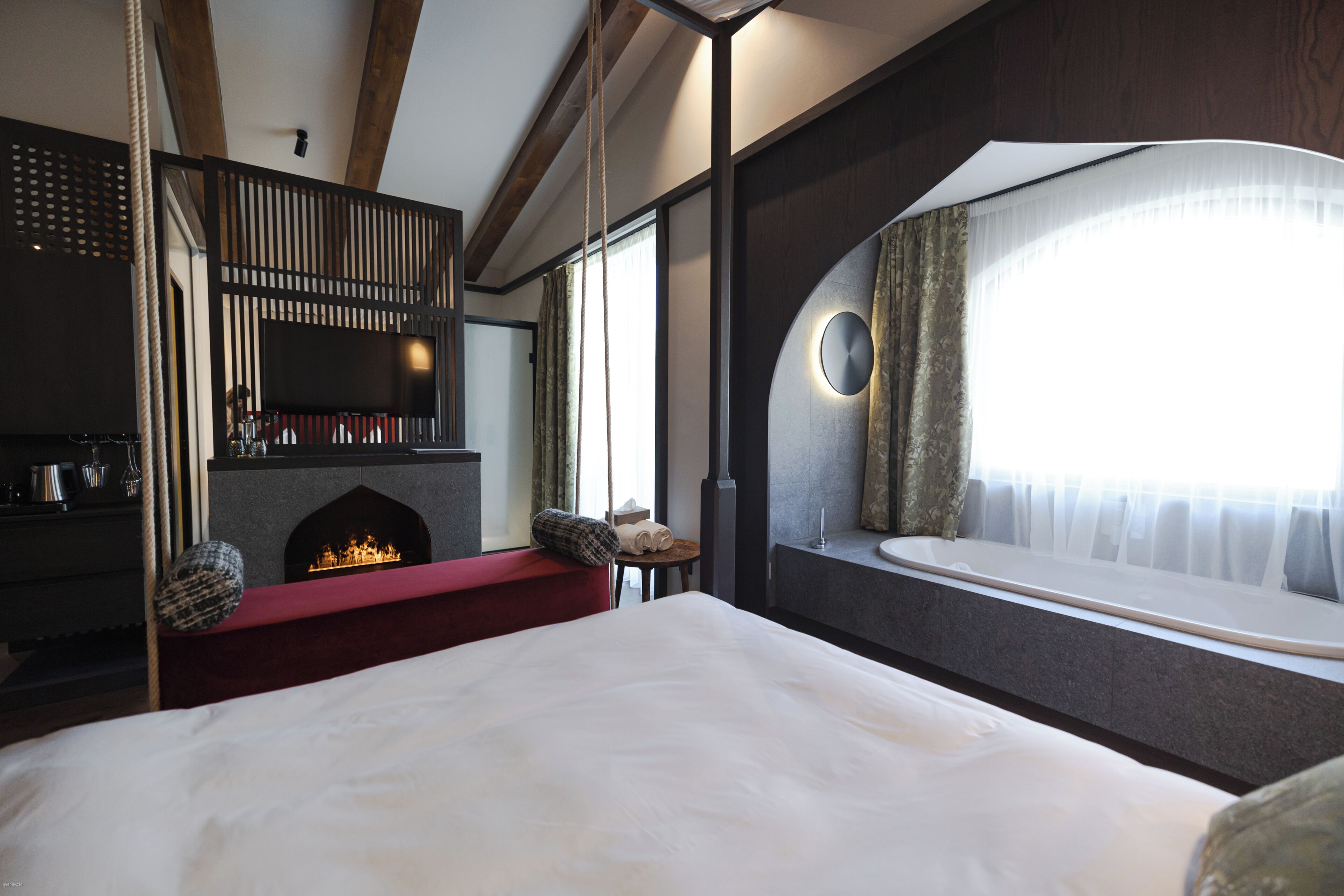 Una habitación de hotel con una cama grande, una chimenea, una ventana con cortinas y una pequeña mesa con un banco rojo.