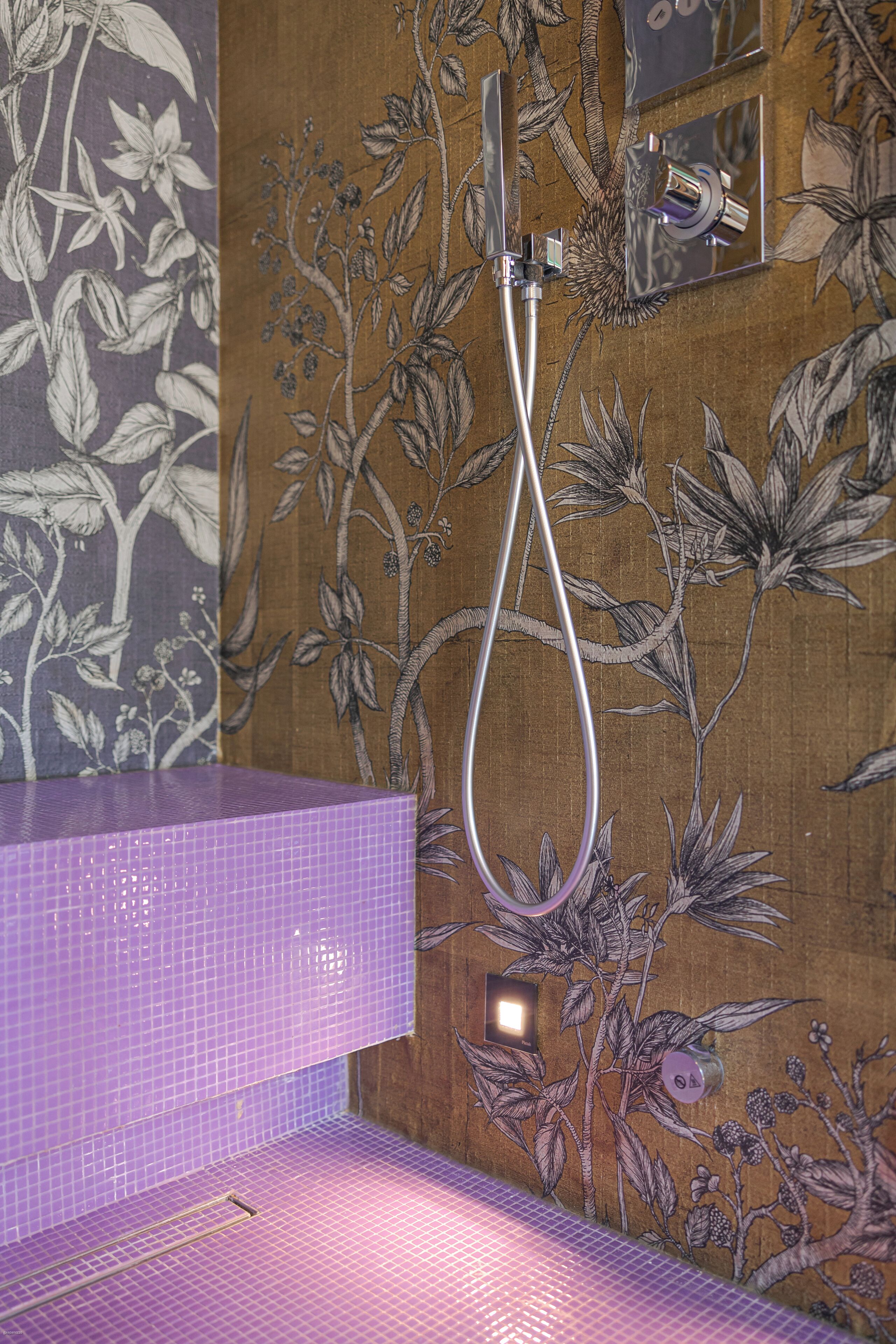 A bathroom with a shower, a purple tiled wall, and a floral wallpaper design.