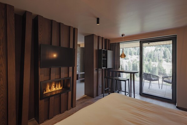 A modern bedroom with a fireplace, a TV mounted on the wall, and a balcony with chairs. - Latemar Hotel Suites & Spa (Soraga)