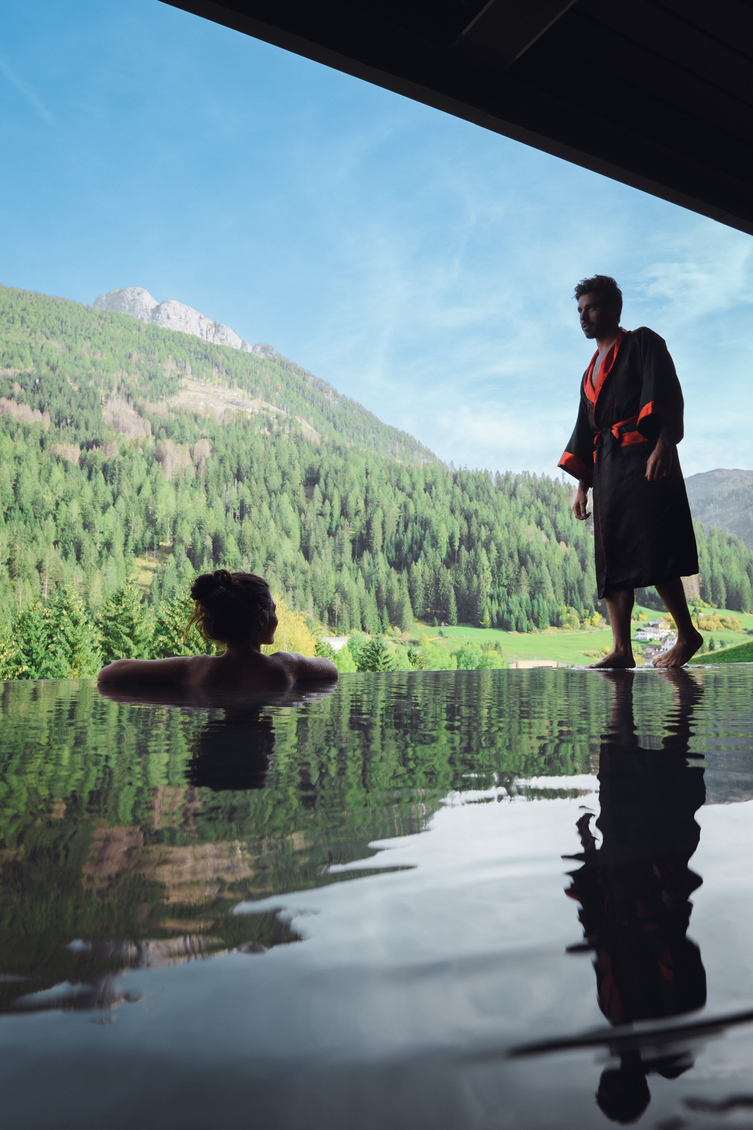Une personne vêtue d’une robe, debout près d’un bassin aux reflets argentés, avec un paysage montagneux en arrière-plan.