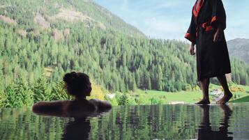 Une personne vêtue d’une robe, debout près d’un bassin aux reflets argentés, avec un paysage montagneux en arrière-plan.