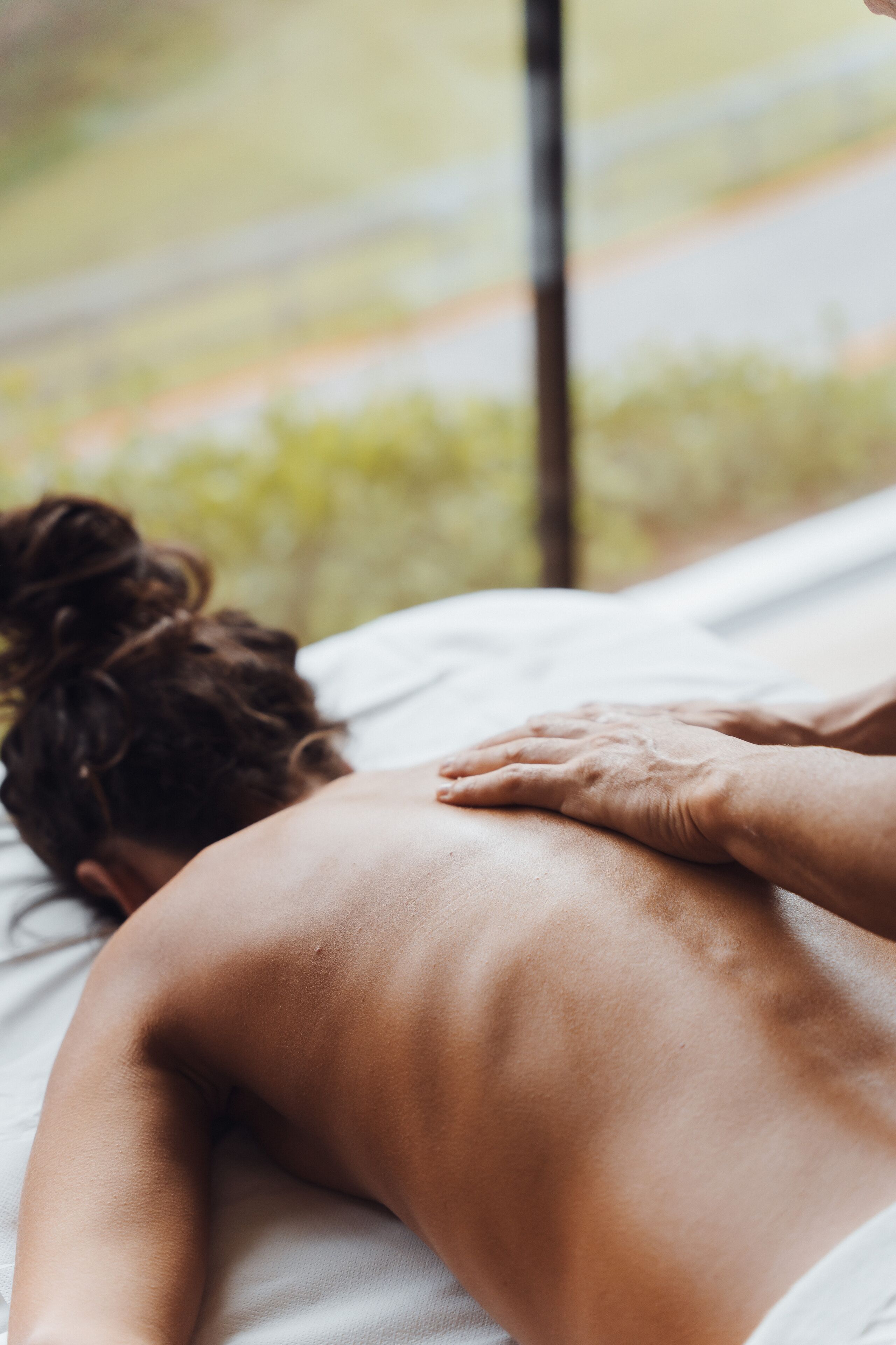 A person receiving a back massage on a bed with a view of greenery outside.