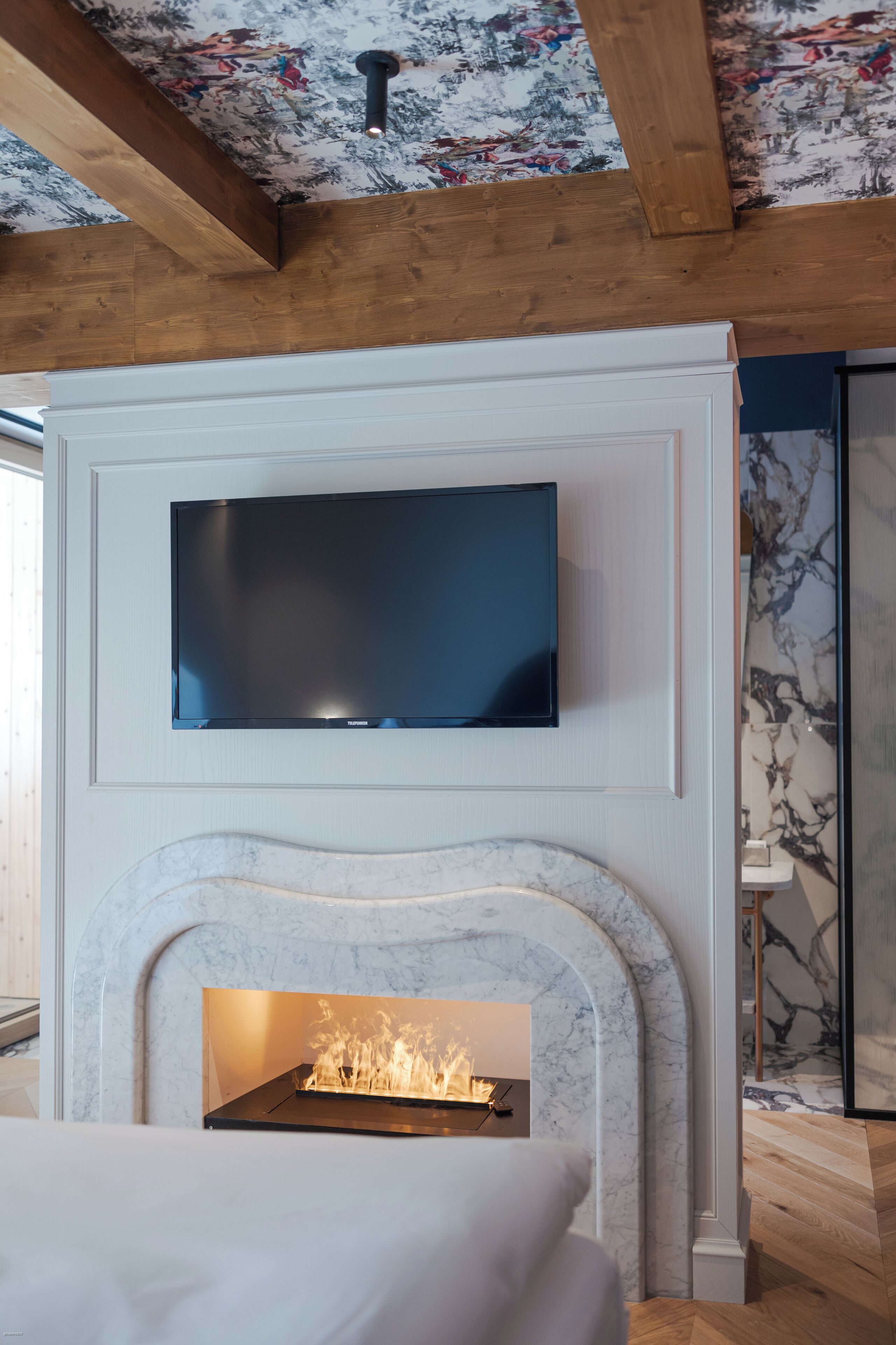 A modern room with a flat-screen TV mounted above a marble fireplace, wooden beams on the ceiling, and a patterned wallpaper.