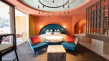 A modern hotel room with a curved ceiling, a large bed with a blue headboard, a bathtub, and a seating area with a blue sofa and ottoman.