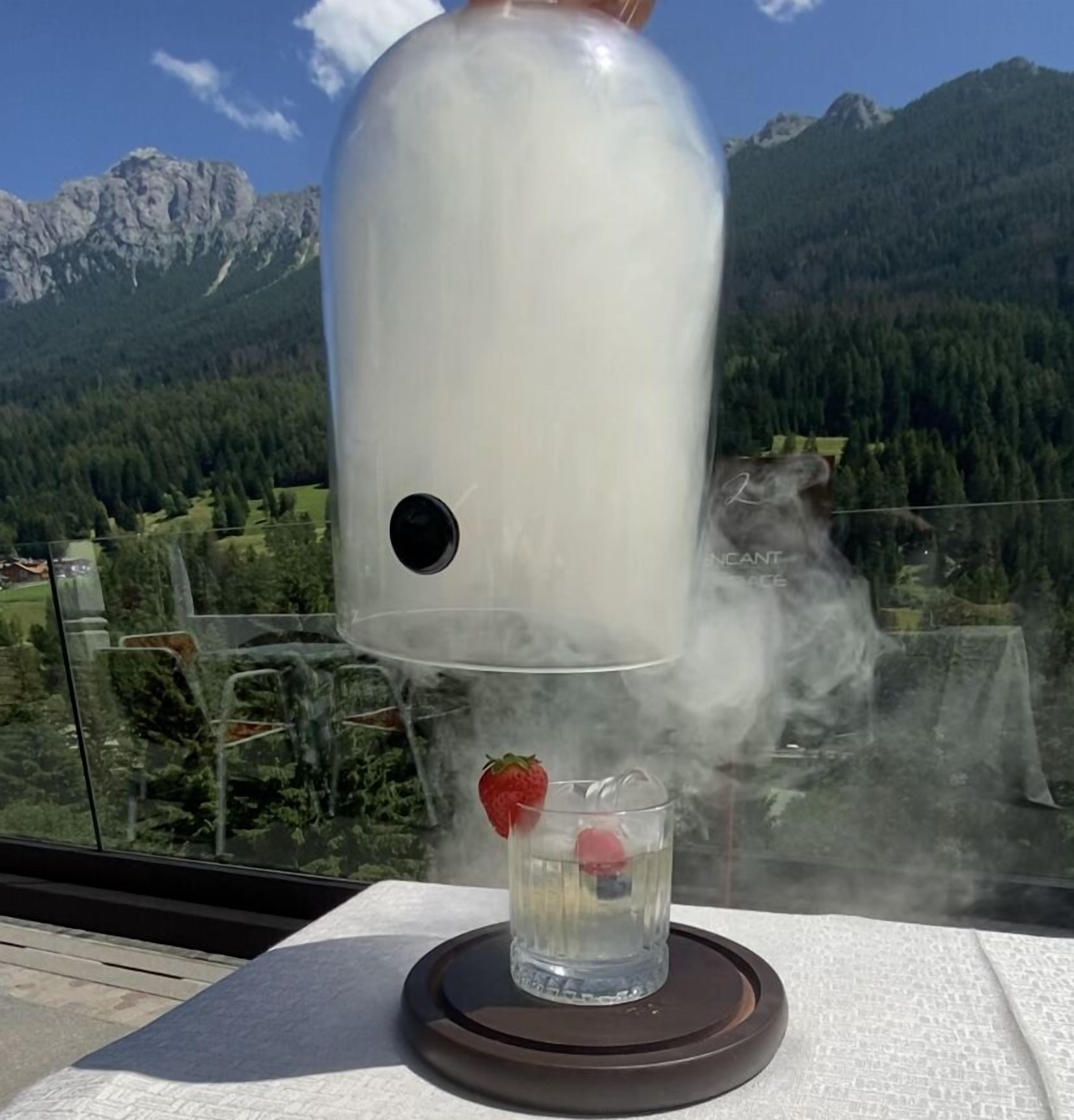 A clear glass of water with a strawberry on a coaster, a white teapot, and a scenic mountain view in the background.