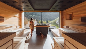 A sauna with wooden benches and a central steam room, large windows offering a view of a forested landscape. - Latemar Hotel Suites & Spa (Soraga)