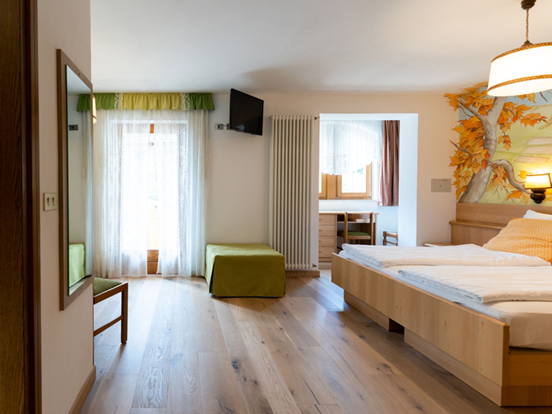 A hotel room with a wooden floor, a bed with a white mattress, a green bench, a desk with a chair, a TV on the wall, and a window with curtains.