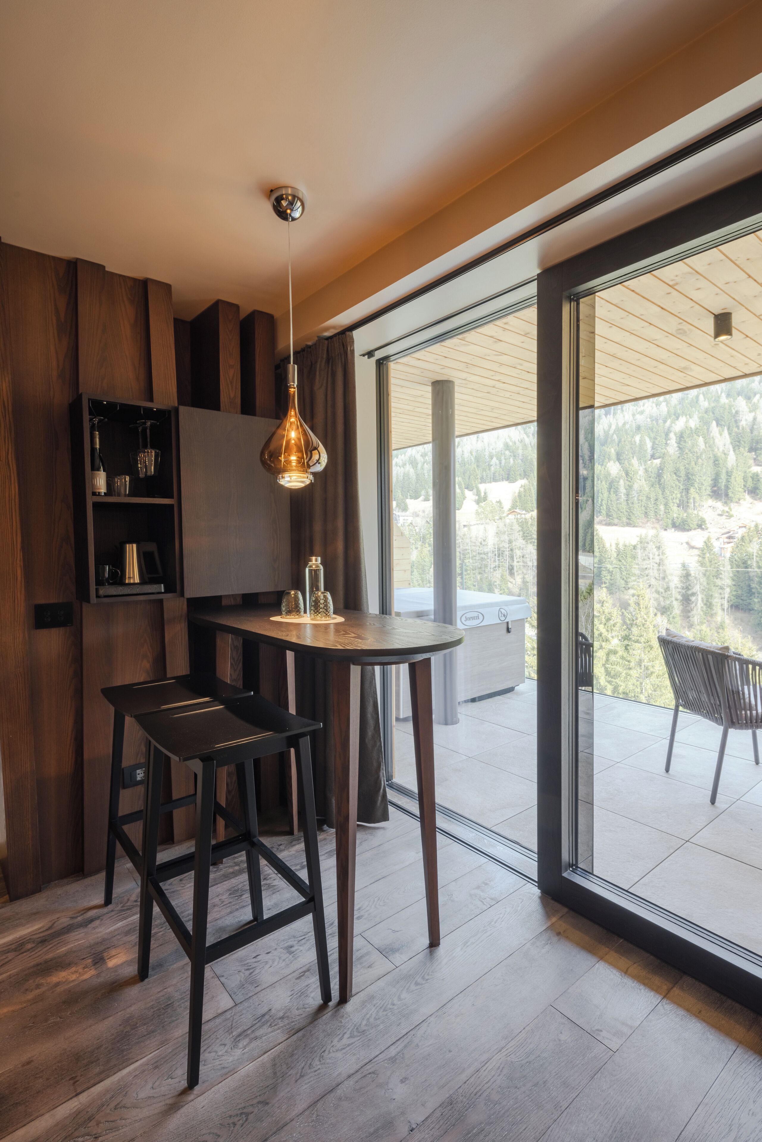 A modern kitchen with a wooden interior, a round dining table, and a large sliding glass door leading to a balcony with a forest view.
