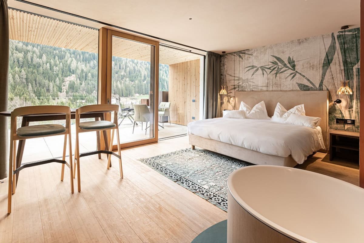 Camera moderna con un letto grande, un angolo salotto con poltrone, una vasca da bagno e vista su una foresta attraverso le porte vetrate.