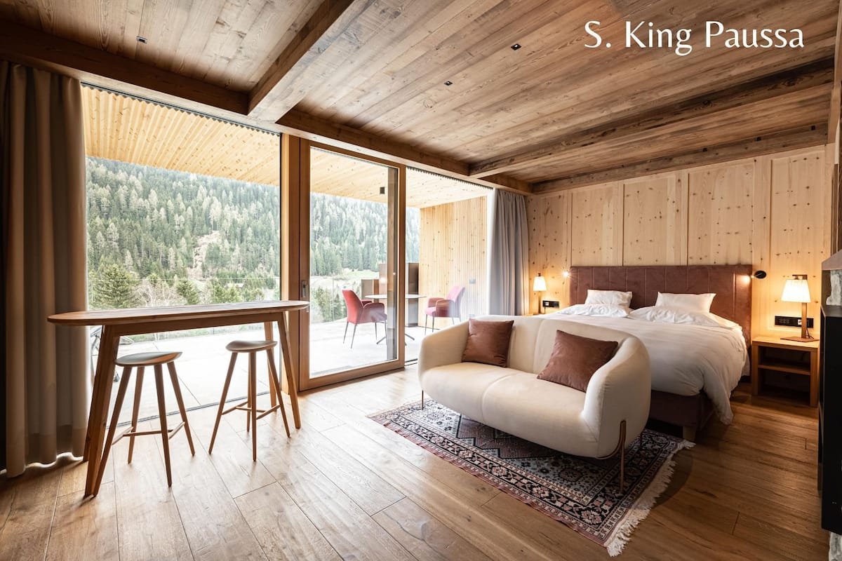 Camera da letto moderna con un letto grande, soffitto in legno e vista su una zona boschiva.