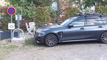 Un coche familiar gris estacionado sobre grava, con un cartel de prohibido estacionar, una estación de carga y una silla blanca al fondo.
