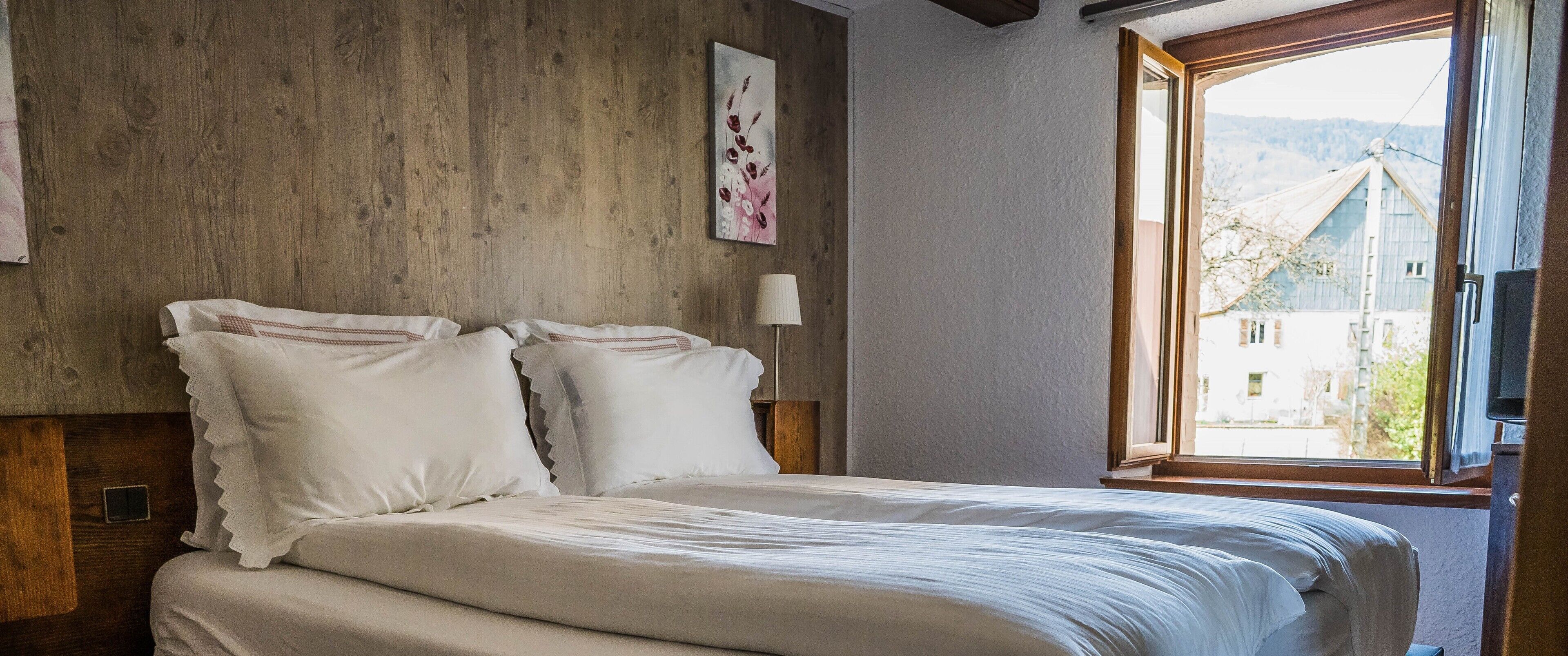 A bedroom with a wooden headboard, white bedding, a bedside lamp, and a window with a view of a building and mountains.