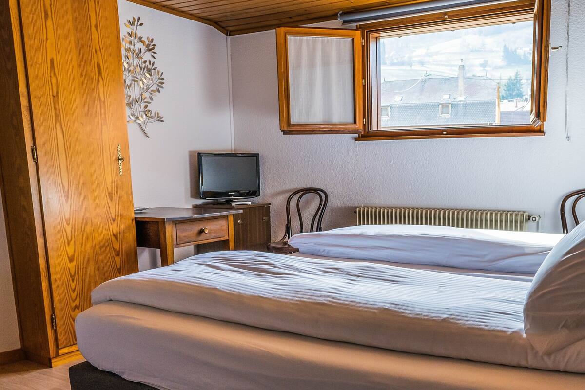 Un dormitorio con cama, mesita de noche, televisor, ventana y lámpara.