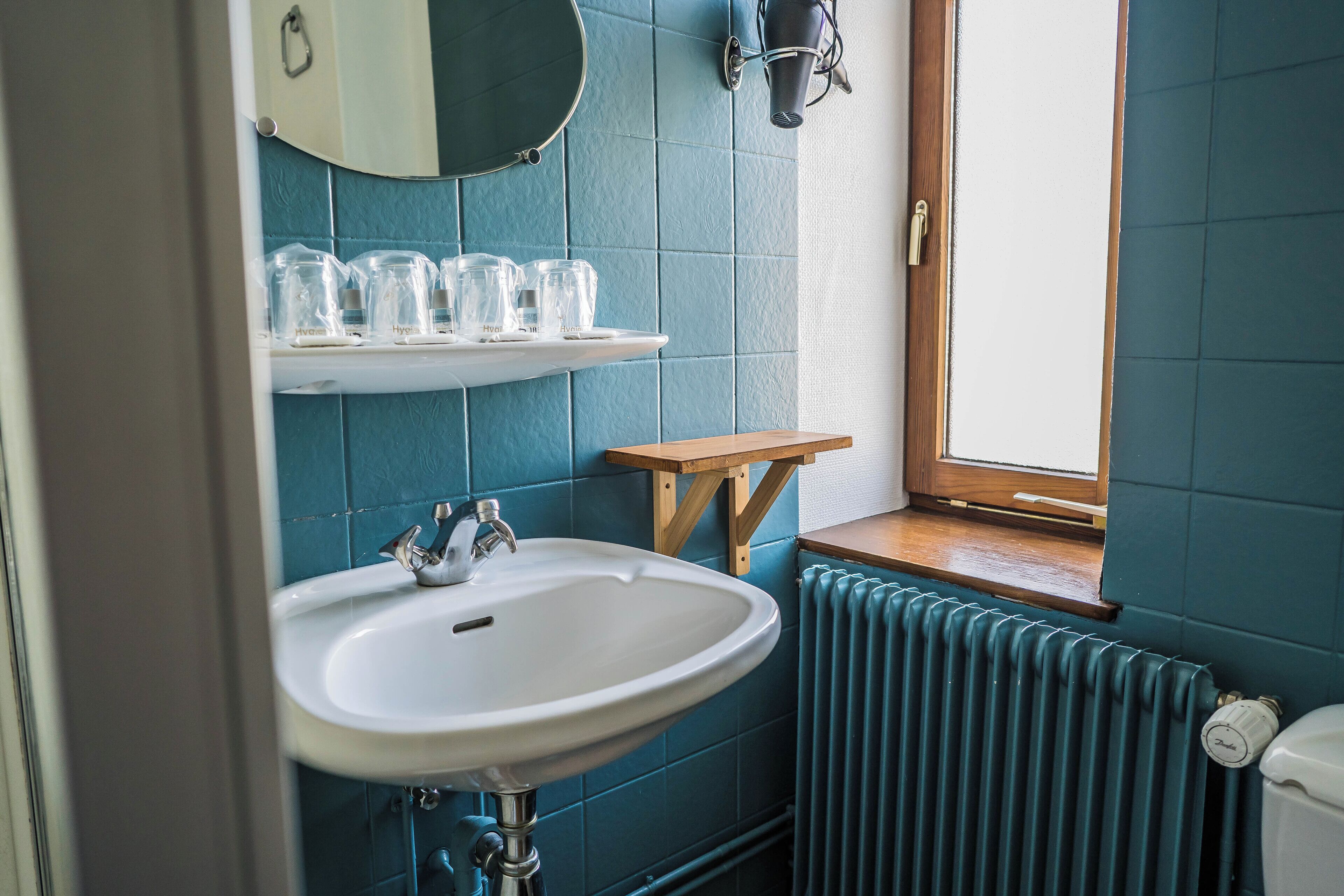 Une salle de bain avec un lavabo blanc, un miroir au-dessus et une étagère avec des verres. Il y a une fenêtre avec un cadre en bois et un radiateur en dessous.