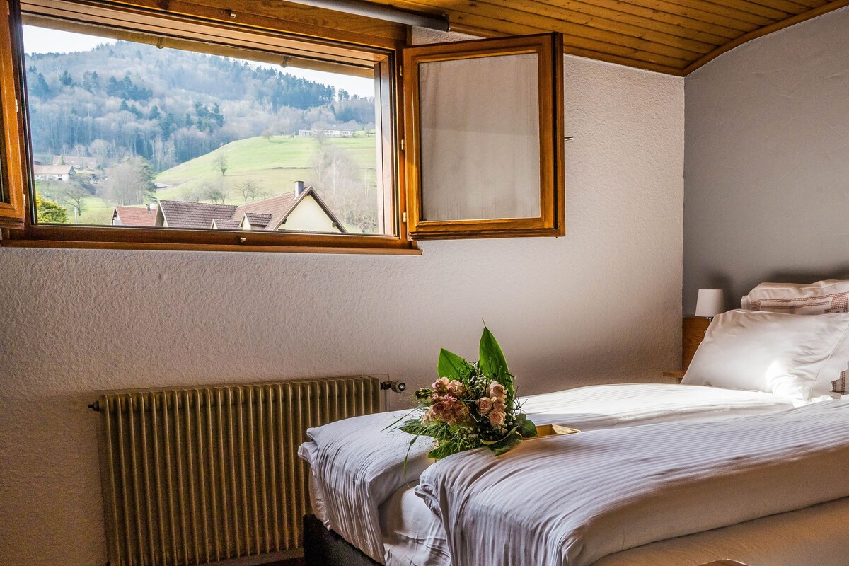Une chambre avec une grande fenêtre, un radiateur, un lit recouvert de draps blancs et un bouquet de fleurs posé sur le lit.