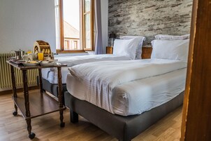 Quarto de hotel com duas camas, uma mesa de madeira com itens de café da manhã, um radiador e uma janela grande com cortinas.
