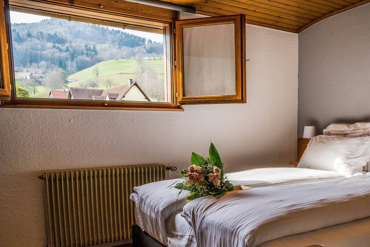 Un dormitorio con un ventanal grande, un radiador, una cama con ropa blanca y un arreglo floral sobre la cama.